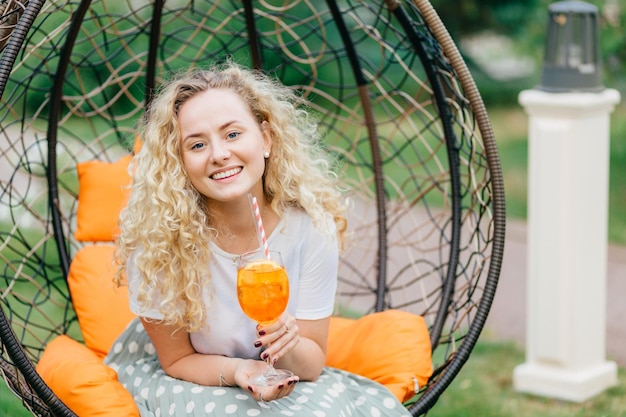 Adorável jovem fêmea encaracolada com cabelo espesso encaracolado segura copo de coquetel laranja posa na cadeira ao ar livre pendurada expressa positividade tem uma conversa agradável com o interlocutor Conceito de lazer