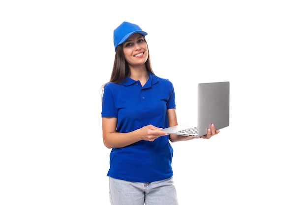Adorável jovem morena caucasiana em camiseta uniforme e boné segurando laptop em fundo branco
