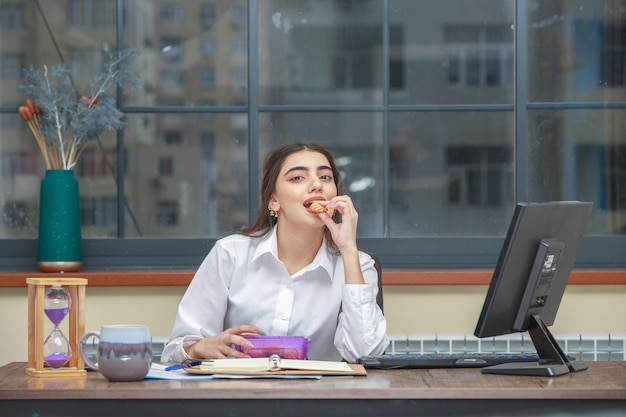 Adorável linda garota comendo cooke no escritório e olhando para a câmera Foto de alta qualidade