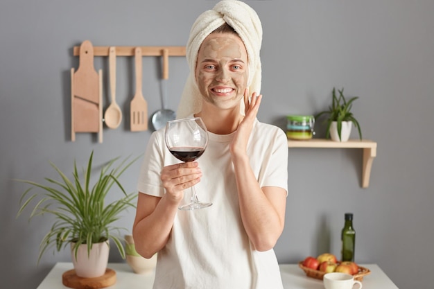 Adorável linda jovem caucasiana feliz posando na cozinha com toalha na cabeça fazendo procedimentos de beleza de cuidados com a pele e segurando um copo de vinho aproveitando o tempo relaxado