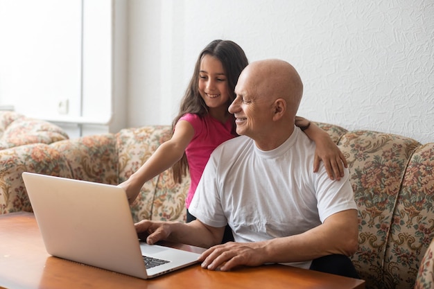 Adorável menina abraçando feliz avô usando laptop em casa.