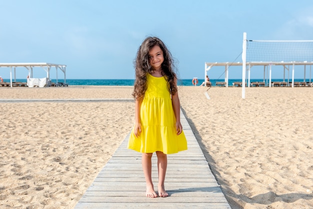 Adorável menina cacheada com um vestido amarelo brincando na praia de areia branca