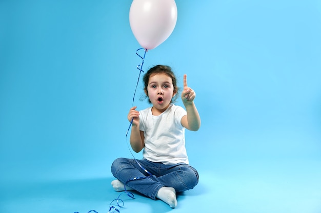 Adorável menina com um balão rosa na mão apontando o dedo em uma superfície azul com espaço de cópia para promoção e publicidade