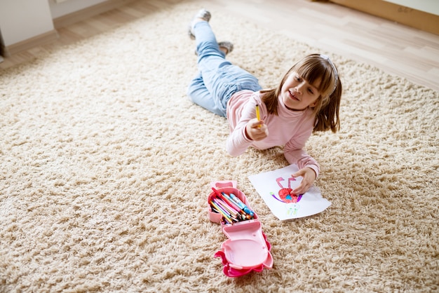 Foto adorável menina criança bonita desenho em papel com giz de cera de madeira enquanto estava deitado no tapete no quarto dela.