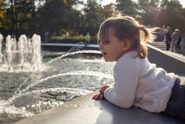 Adorável menina criança olha para a lagoa com fontes no parque em um dia ensolarado passeio em família de fim de semana passando tempo com foco artístico de crianças