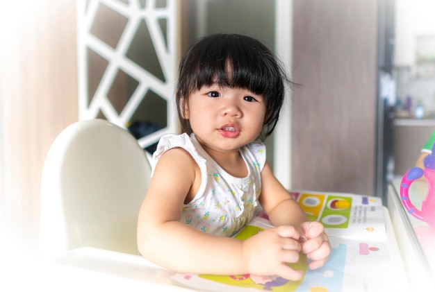 Adorável menina de 18 meses sentada na cadeira de bebê e lendo livro em casa