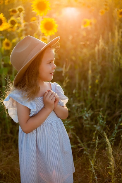 Adorável menina de 4 anos com cabelo loiro comprido encaracolado em um vestido azul e chapéu em um campo de girassol Retrato de verão de uma criança feliz Pôr do sol