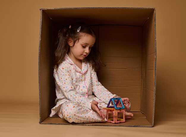 Adorável menina de pijama sentada dentro de uma caixa de papelão e brincando com uma pequena casa colorida construída a partir de blocos de construção magnéticos