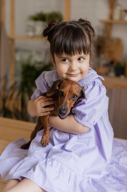 Adorável menina em um lindo vestido brinca com um cachorro dachshund na cozinha e a alimenta