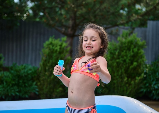 Adorável menina em um maiô se divertindo soprando bolas de sabão em uma piscina inflável no jardim em casa