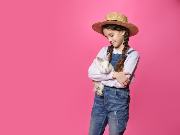 Adorável menina encaracolada de chapéu de palha segurando um coelhinho fofo olhando para a câmera isolada fundo rosa