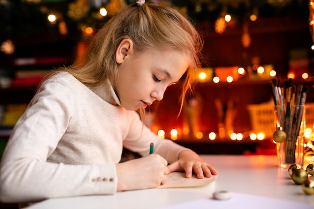 Adorável menina está escrevendo uma carta para Santa Klaus sentado em cima da mesa