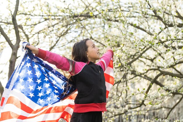 Adorável menina feliz sorrindo e acenando a bandeira americana. Feriado patriótico. Criança feliz, menina bonitinha com bandeira americana. EUA comemoram 4 de julho. Conceito do dia da independência.