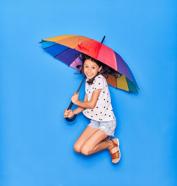 Adorável menina hispânica sorrindo feliz segurando guarda-chuva colorido pulando com sorriso no rosto sobre fundo azul isolado