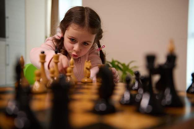 Adorável menina pegando uma peça de xadrez enquanto jogava xadrez em casa. Jogos de tabuleiro intelectuais inteligentes para crianças, desenvolvimento lógico, conceito de educação