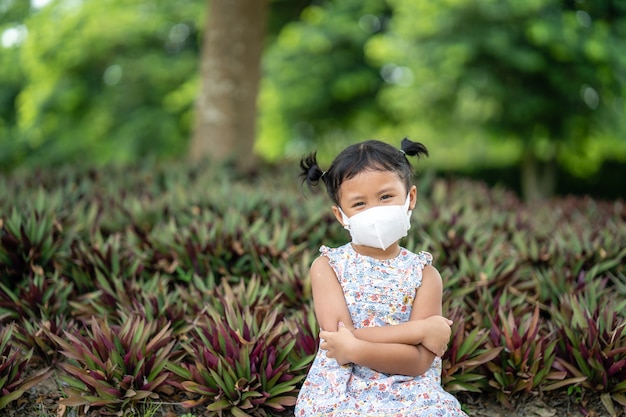 Adorável menina pequena com máscara facial saudável sentada contra uma planta no parque