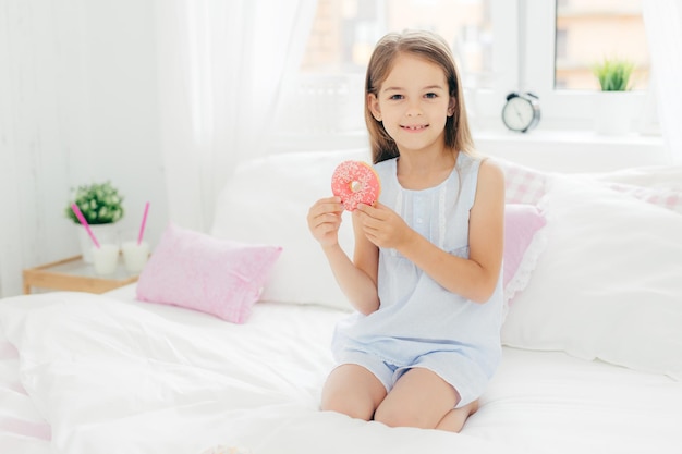 Adorável menina pequena segura rosquinha deliciosa nas mãos vai tomar café da manhã posa na cama no quarto branco aconchegante Olha para a câmera com expressão positiva Conceito de hora de dormir de sobremesa de crianças