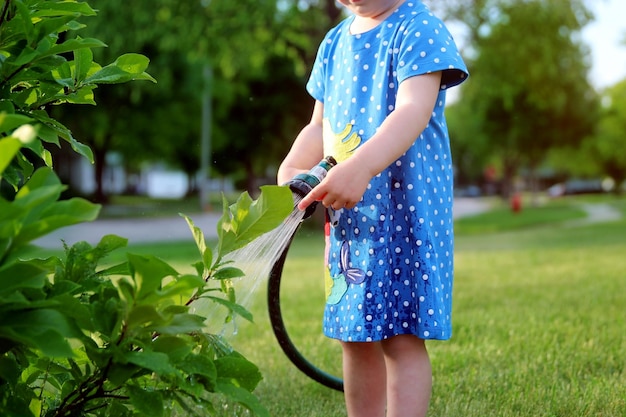 Adorável menina regando a árvore com uma mangueira de jardim na noite quente e ensolarada de verão