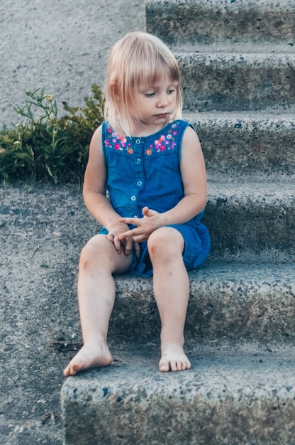 Adorável menina rindo da natureza - garota feliz em um vestido azul e espancada pelos joelhos senta-se na escada. infância feliz.