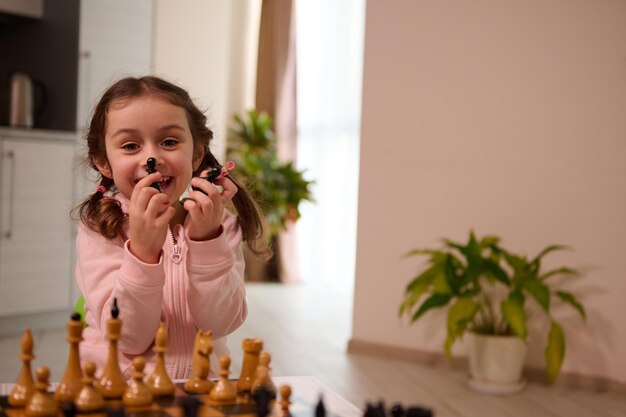 Adorável menina segurando peças de xadrez e sorrisos fofos com alegre sorriso cheio de dentes olhando para a câmera enquanto jogava xadrez com seu irmão no interior da casa. Jogo de xadrez para mente inteligente
