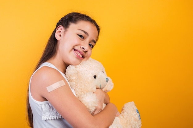 Adorável menina segurando um ursinho de pelúcia com um curativo no braço.