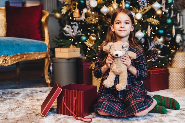 Adorável menina senta-se no chão perto de embrulhos de árvores decoradas de Ano Novo presentes sendo feliz em receber ursinho de Papai Noel Pequena criança fofa recebe presente no conceito de milagre e feriados de Natal