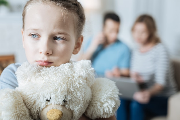 Adorável menina triste de olhos azuis abraçando seu ursinho de pelúcia e se sentindo sozinha enquanto seus pais trabalhavam em seus laptops