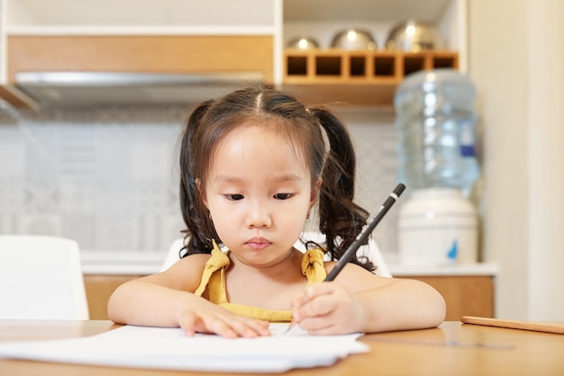 Adorável menina vietnamita sentada à mesa da cozinha desenhando a lápis