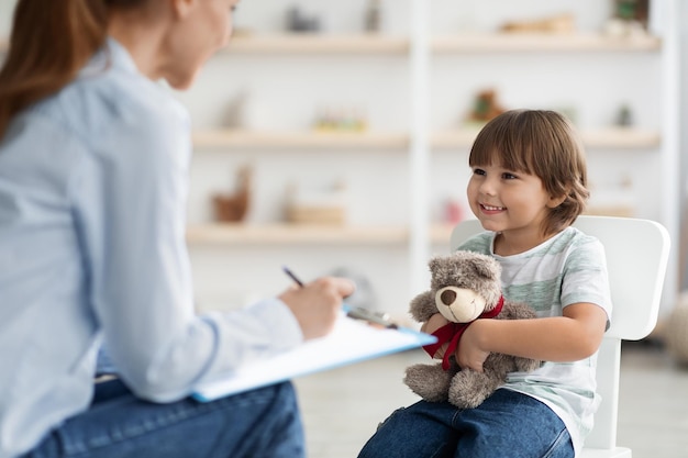 Adorável menino abraçando ursinho de pelúcia e sorrindo para especialista sentado no escritório de psicoterapeuta