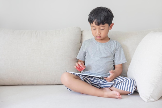 Adorável menino asiático feliz vestindo uma camisa cinza e shorts listrado azul-branco está se divertindo brincando com seu tablet em um sofá creme olhando para a tela do celular