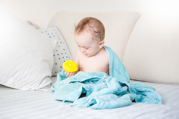 Adorável menino coberto de toalha azul brincando com pato de borracha amarelo no sofá depois do banho