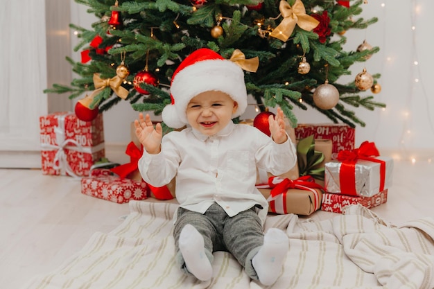 Adorável menino com chapéu de ano novo se senta em um cobertor na frente da árvore de natal.