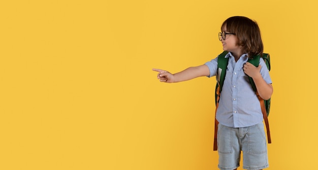 Adorável menino de óculos com mochila apontando de lado para o espaço vazio e sorrindo panorama de fundo laranja