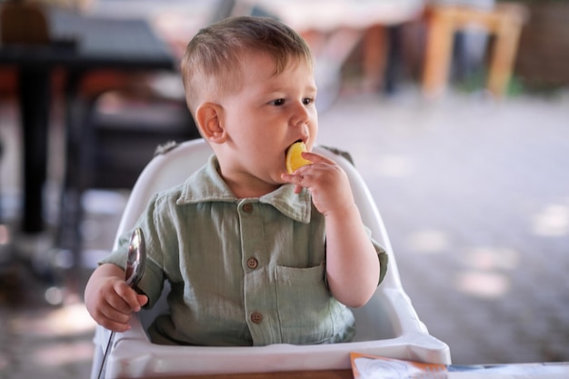 Adorável menino lambendo pedaço de fruta limão sentado na cadeira alta e segurando a colher no jantar