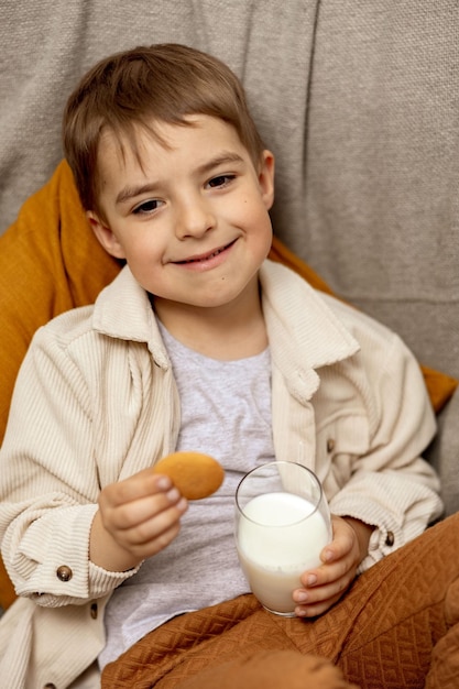 Adorável menino sentado no sofá em casa bebendo leite com biscoito Leite fresco em vidro laticínios bebida saudável Fonte de saúde de lactose cálcio Criança pré-escolar com roupas casuais