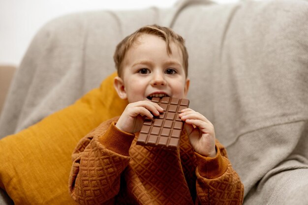 Adorável menino sentado no sofá em casa e comendo barra de chocolate Criança e doces de confeitaria Criança desfrutar de uma deliciosa sobremesa Criança pré-escolar com roupas casuais Emoção positiva