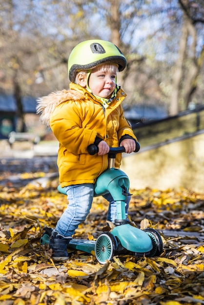 Adorável menino usando capacete protetor amarelo montando scooter de bebê ao ar livre no dia de outono