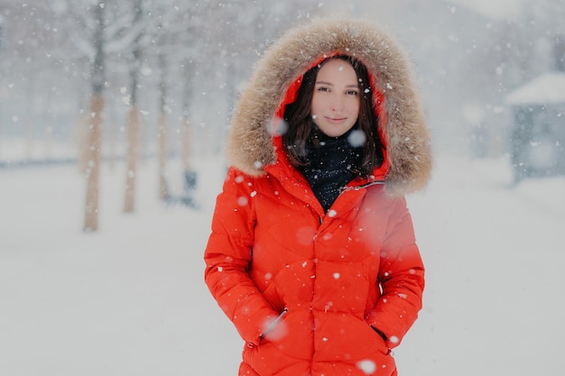 Adorável modelo feminino de jaqueta vermelha fica ao ar livre durante a queda de neve olha com olhos escuros para a câmera vai passear com o namorado respira ar fresco Pessoas inverno e conceito de tempo livre