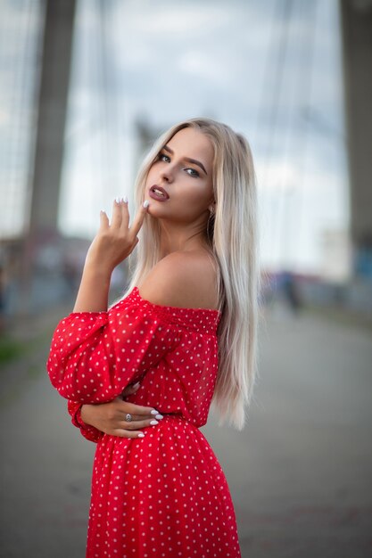 Adorável modelo loira com um vestido vermelho da moda, posando na ponte à noite