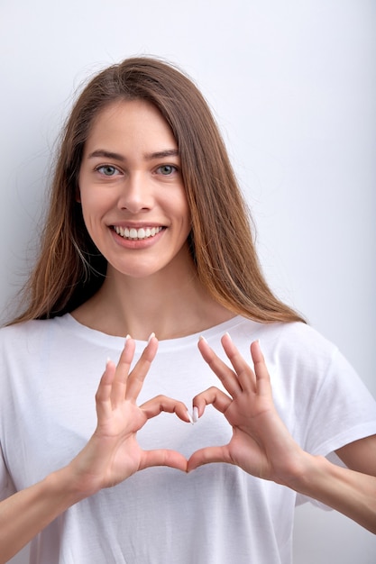 Adorável mulher bonita mostrando coração gesto de amor sorrindo posando para a câmera isolada no estúdio branco bac ...