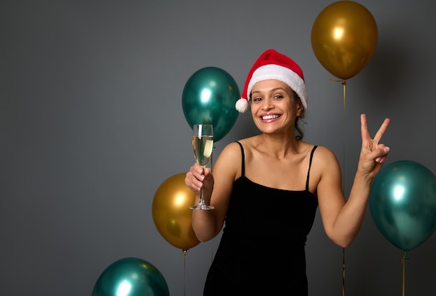 Adorável mulher com um chapéu de Papai Noel segura uma flauta com champanhe e mostra o símbolo da paz olhando para a câmera contra a decoração de Natal com balões de ar brilhantes de luxo no fundo cinza da parede com espaço para anúncio de cópia