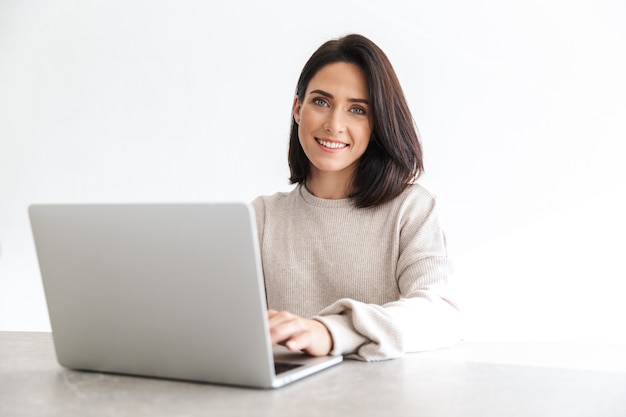 adorável mulher de 30 anos trabalhando em um laptop, enquanto está sentada sobre uma parede branca em uma sala iluminada