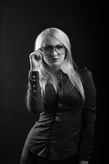 Adorável mulher loira com cabelo comprido usa óculos e blusa, posando no estúdio. Foto monocromática