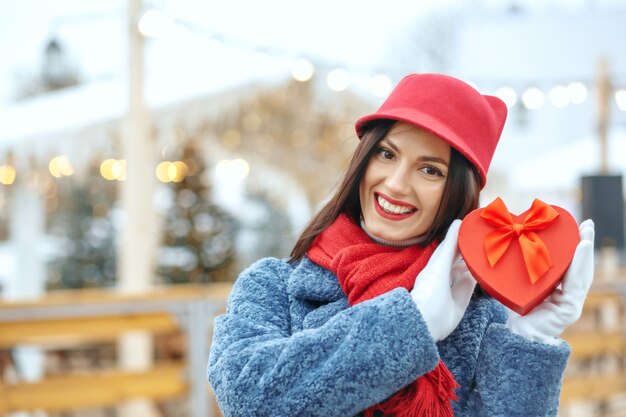 Adorável mulher morena com casaco de inverno, segurando uma caixa de presente na feira de Natal. Espaço para texto