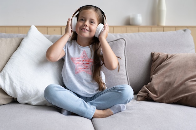 Adorável pequena colegial sentada no sofá, usando modernos fones de ouvido sem fio, ouvindo sua música favorita, Linda criança descansando no sofá