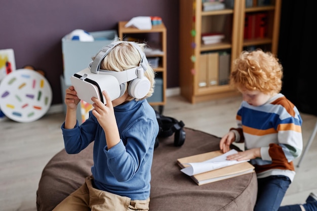 Adorável pequeno aluno da creche em fone de ouvido vr jogando jogo virtual enquanto está sentado na frente