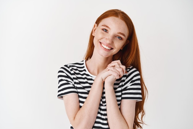 Adorável ruiva admira algo apertando as mãos juntas e sorrindo agradecido em pé encantado contra o fundo branco