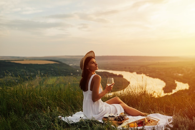 Foto adorável tenra garota vestida de branco com chapéu de palha, fazendo um piquenique no pôr do sol de verão na montanha.