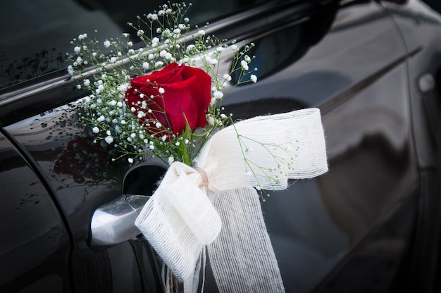 El adorno de rosa roja con lazo blanco decora muy bien el asa plateada del coche de boda negro. Concepto de detalle de ceremonia