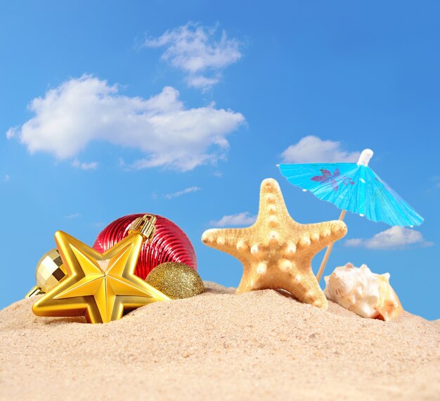 Adornos navideños, conchas y estrellas de mar en la arena de una playa contra el cielo azul